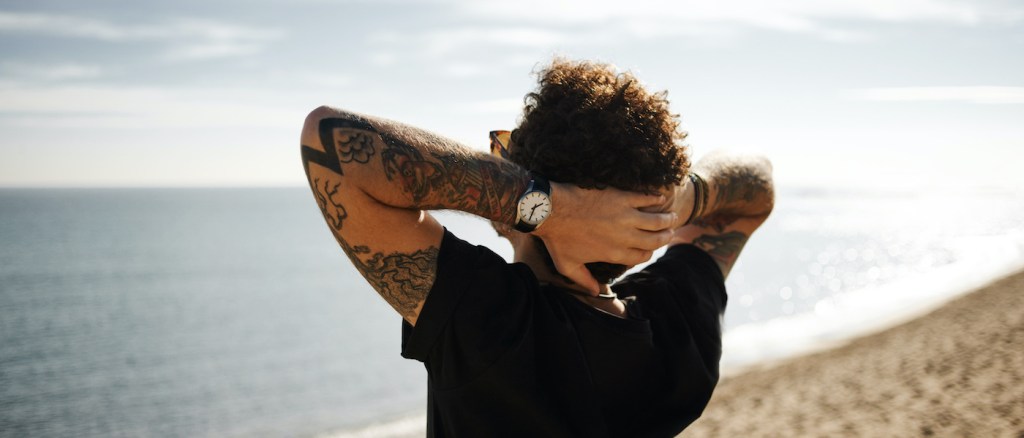 Ritratto ravvicinato di un uomo con la schiena rivolta verso la spiaggia, in piedi sulla sabbia, con le mani dietro la testa e lo sguardo rivolto in lontananza, mentre si gode il sole