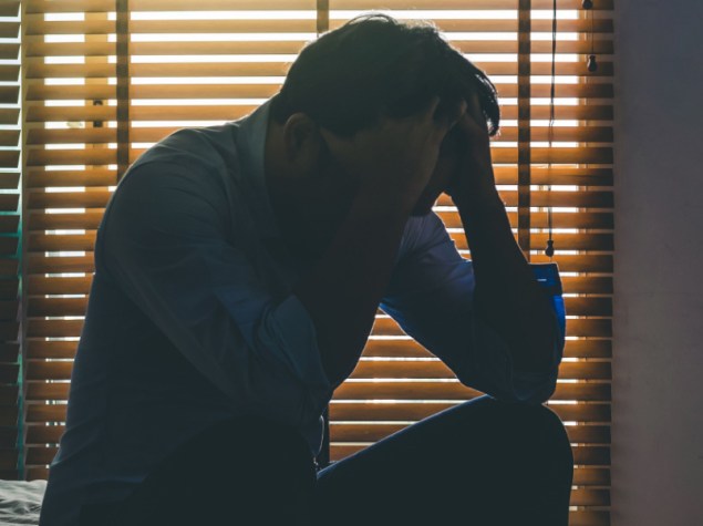 Uomo depresso seduto con la testa tra le mani sul letto nella camera da letto buia con ambiente a bassa luminosità, concetto drammatico