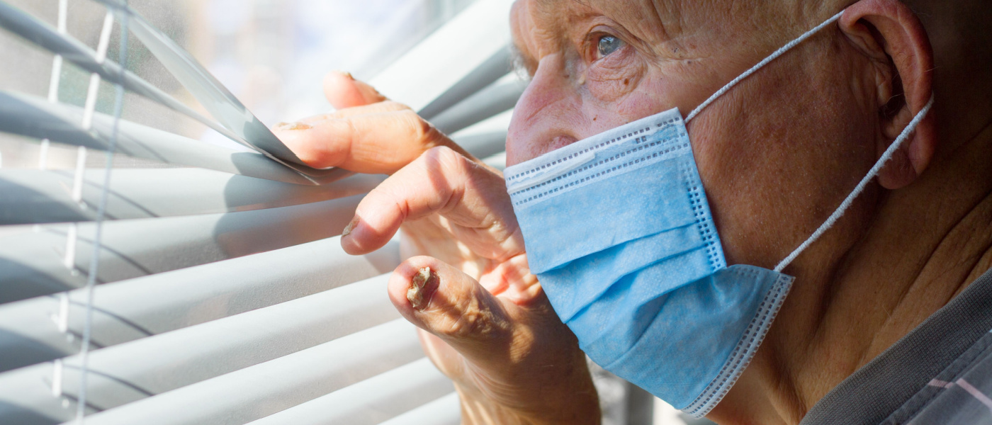 Un uomo molto anziano con una maschera protettiva blu sul viso guarda fuori dalla finestra, concetto di "stay at home".