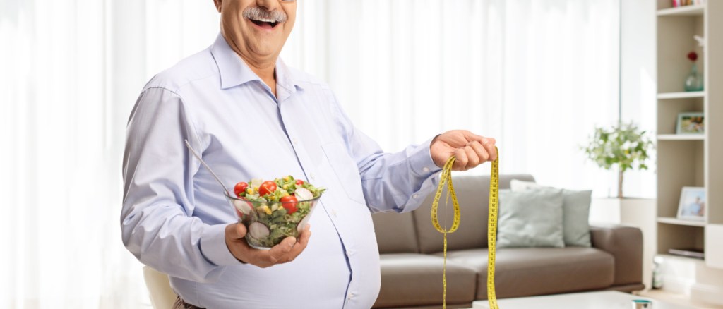 Uomo maturo con in mano un'insalata e un nastro di misurazione all'interno di un soggiorno