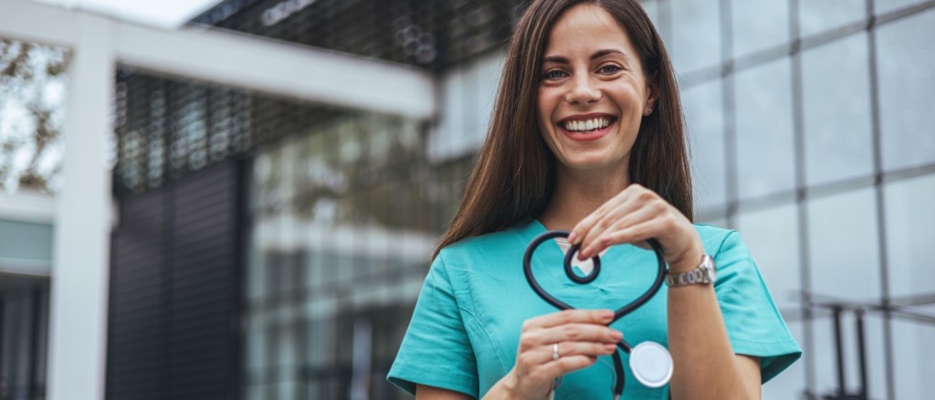 A joyful Caucasian female nurse forms a heart with her stethoscope, displaying care outside a medical facility, dressed in scrubs, symbolizing compassion in healthcare.