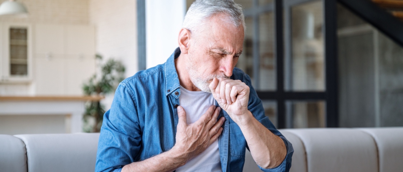 Uomo anziano seduto sul divano a casa con una mano sul petto. L'uomo ha un attacco d'asma, difficoltà respiratorie, forte dolore o dispnea.