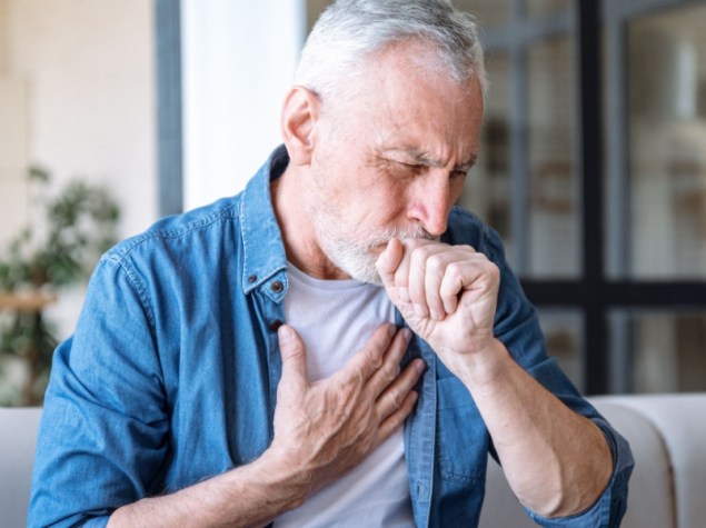 Uomo anziano seduto sul divano a casa con una mano sul petto. L'uomo ha un attacco d'asma, difficoltà respiratorie, forte dolore o dispnea.