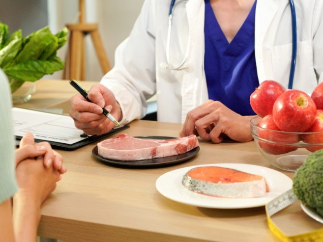 Professionista sanitario durante una consulenza nutrizionale con una paziente, circondato da alimenti sani come frutta, verdura, pesce e carne, simbolo di educazione alimentare e benessere.