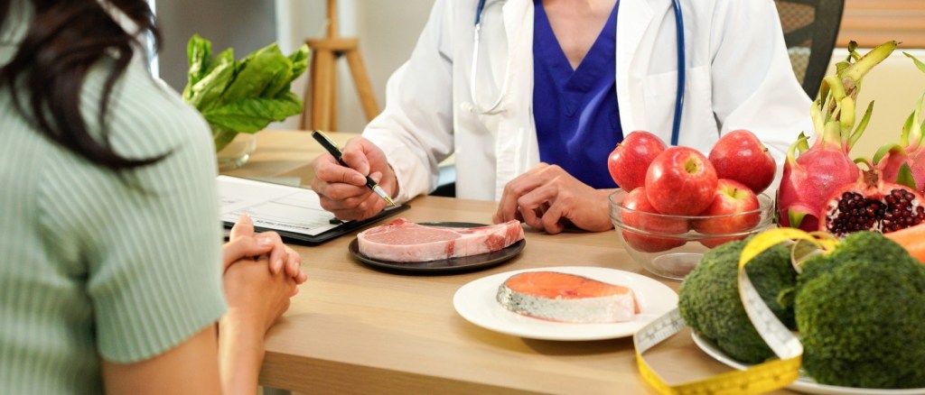 Professionista sanitario durante una consulenza nutrizionale con una paziente, circondato da alimenti sani come frutta, verdura, pesce e carne, simbolo di educazione alimentare e benessere.