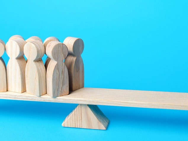 Illustrazione concettuale della disuguaglianza: una bilancia in legno sbilanciata mostra un gruppo di persone da un lato e una singola persona isolata dall’altro, su sfondo azzurro.