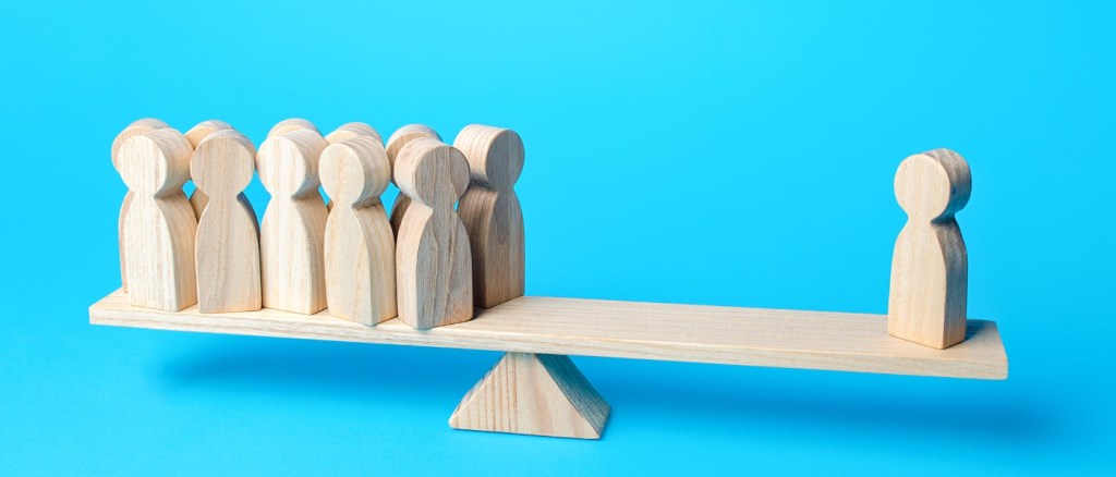 Illustrazione concettuale della disuguaglianza: una bilancia in legno sbilanciata mostra un gruppo di persone da un lato e una singola persona isolata dall’altro, su sfondo azzurro.