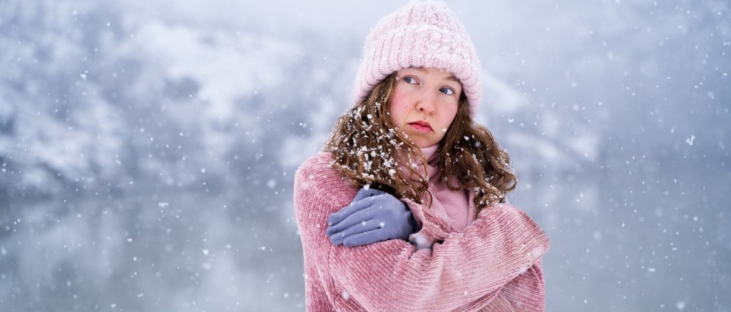 Ritratto di giovane donna fredda che cerca di mantenere caldo all'aperto in inverno, sentendosi freddo in piedi vicino al lago in montagna alla luce del giorno, emozione del tempo freddo e concetto di disagio invernale